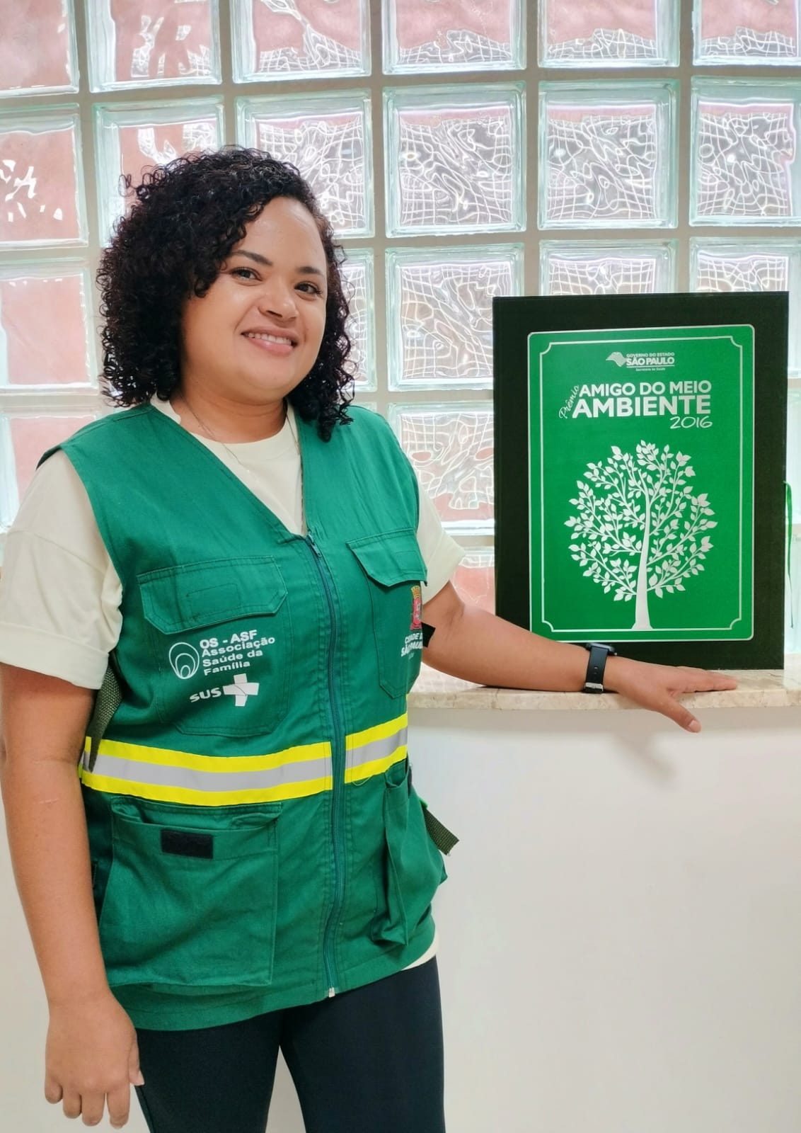 A foto mostra Jane, uma mulher branca, de cabelos pretos e enrolados, usando um colete verde. Ela está ao lado de uma placa do Prêmio Amigo de Meio Ambiente 2016. 